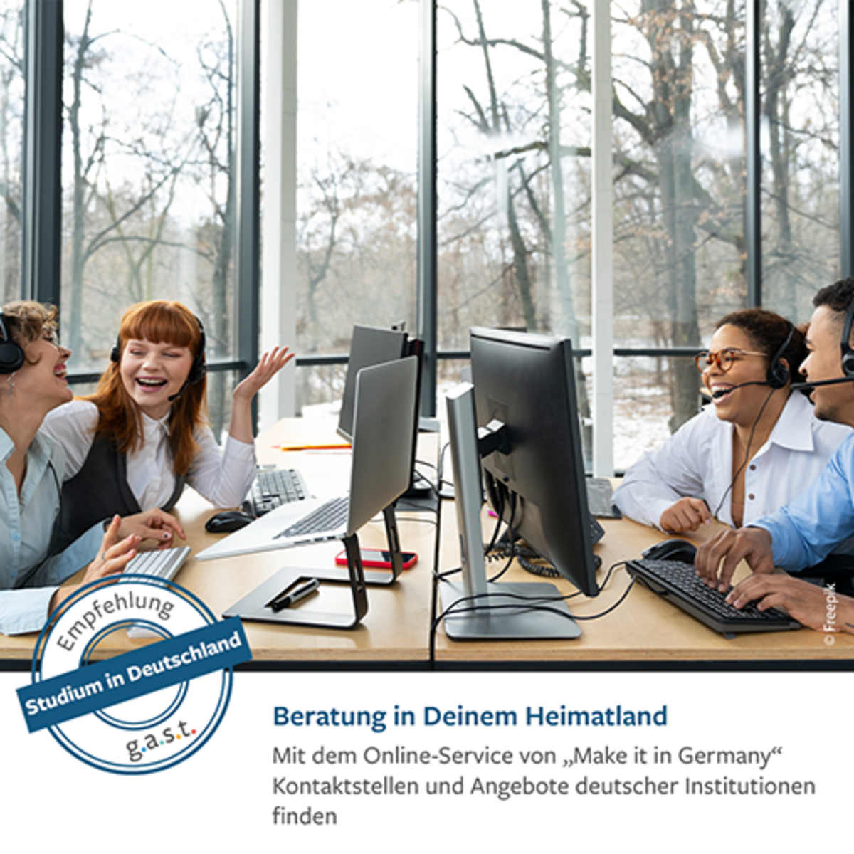 Imagebild zum Thema "Studium in Deutschland": Vier junge Internationals sitzen an einem Tisch vor jeweils einem Bildschirmarbeitsplatz. Sie tragen Hemd bzw. Bluse und lachen. Im Hintergrund öffnet sich vor einer Glasfront der Bild in einen spätherbstlichen winterlichen Wald. Auf dem Waldboden liegt Schnee.