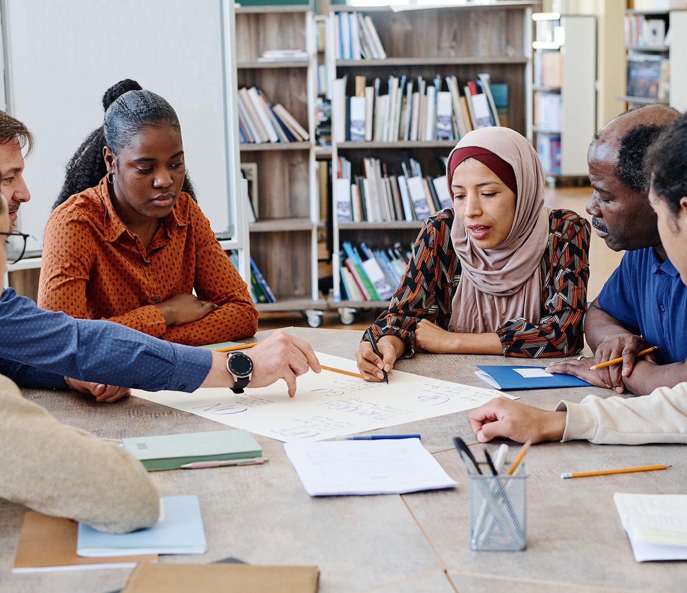Eine Gruppe Internationals mit Lehrkraft sitzt an einem Tisch in einer Bibliothek und arbeitet an einer Aufgabe. 